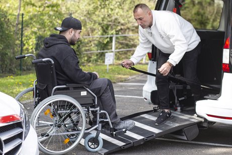 Ein Mitarbeiter der Schwabenfahrdienst sichert einen Mann im Rollstuhl auf einer ausgefahrenen Hebebühne beim Einstieg in ein barrierefreies Fahrzeug.