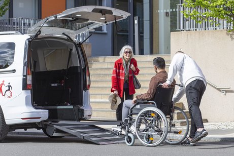 Ein Mitarbeiter der Schwabenfahrdienst unterstützt einen Rollstuhlfahrer beim Einstieg in ein barrierefreies Fahrzeug, während eine ältere Frau daneben steht und zusieht.