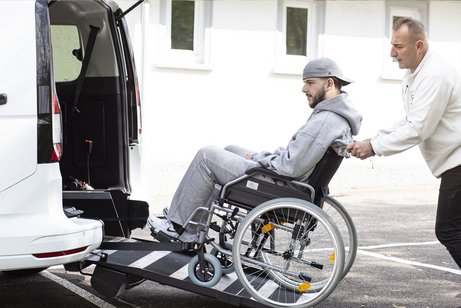Ein Mitarbeiter der Schwabenfahrdienst schiebt einen Rollstuhlfahrer über eine Rampe in ein behindertengerechtes Fahrzeug vor einem Gebäude.
